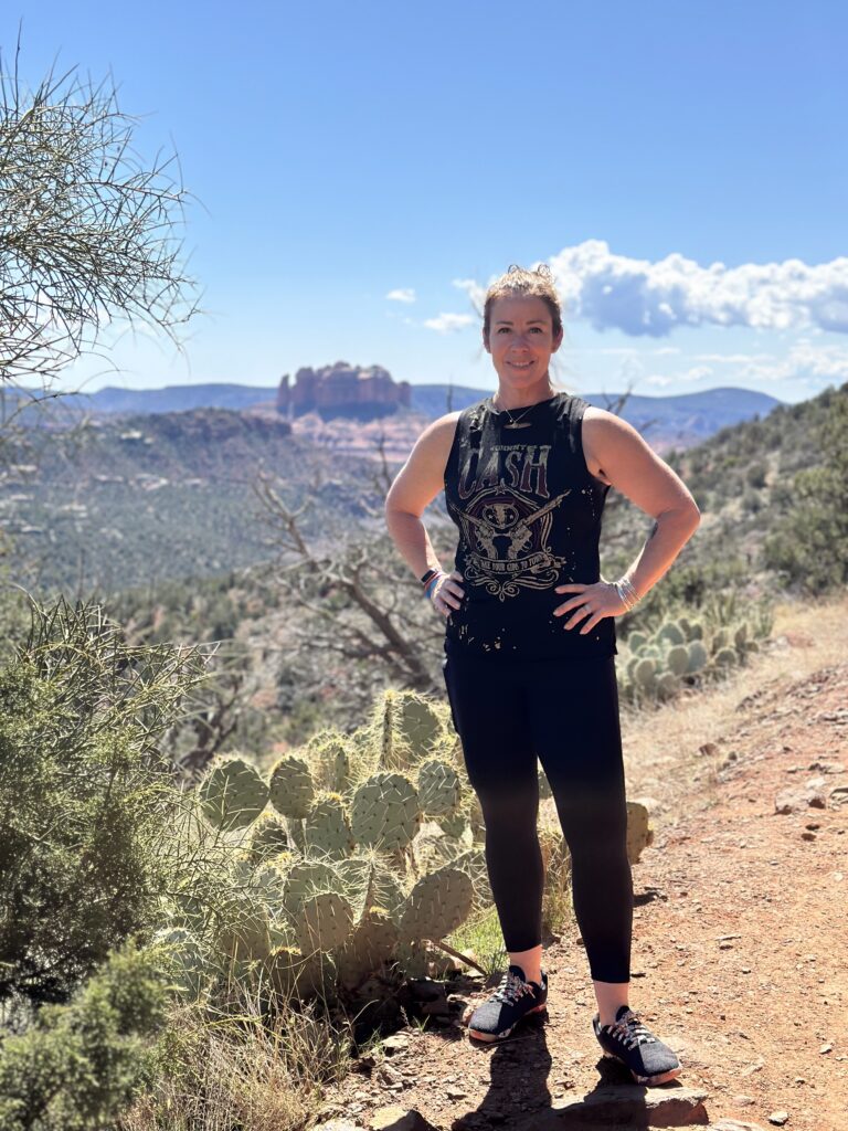 woman standing on a hiking trail in the Sedona desert next to a prickly pear cactus in the sushine, red rock formations in the distance